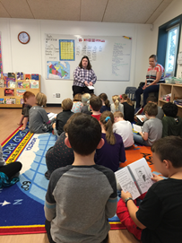 A teacher reads a story to an elementary class with students seated on a carpet.